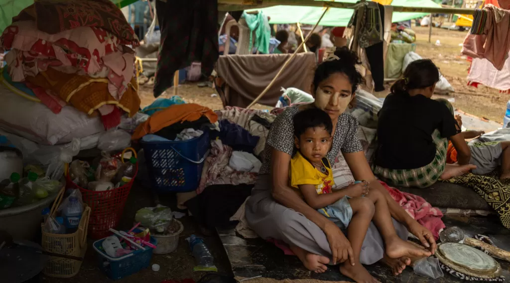 A mother sits on the ground ith her child in her lap in a makeshift shelter, with their belongings around them