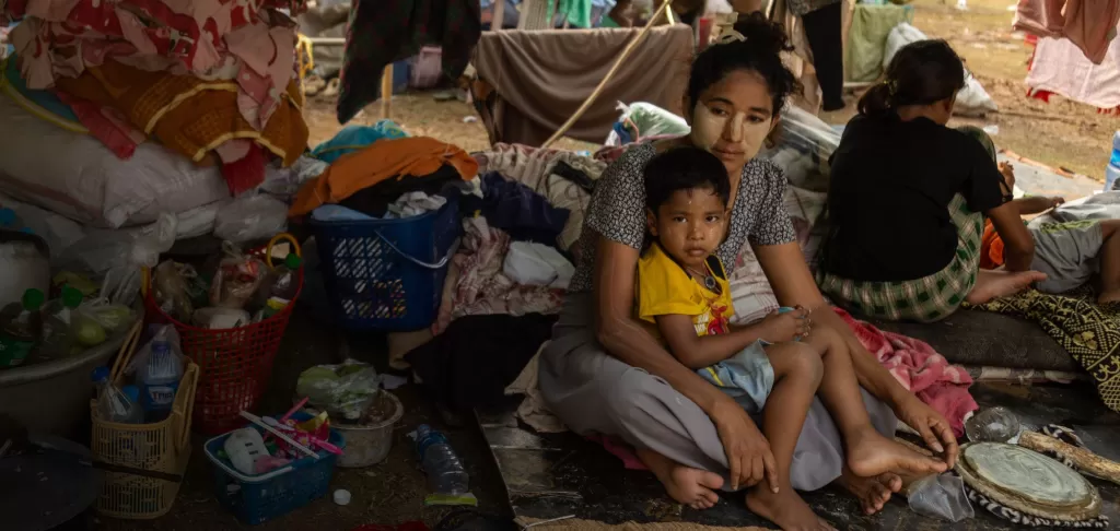 A mother sits on the ground ith her child in her lap in a makeshift shelter, with their belongings around them