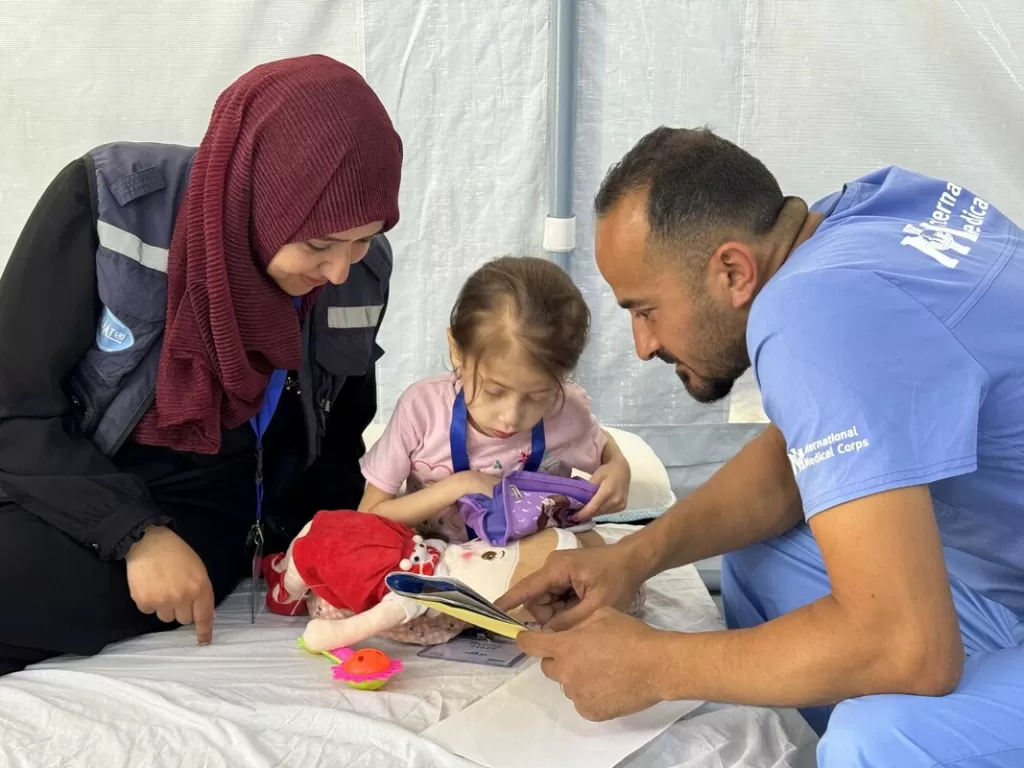 Two humanitarian workers sit with a young girl, engaging her with a book and toys in a medical tent