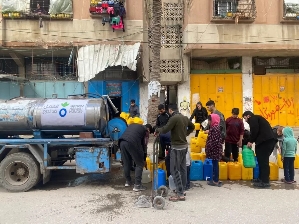 People gathering with yellow and blue water containers around an Action Against Hunger water truck distributing clean water in an urban residential area