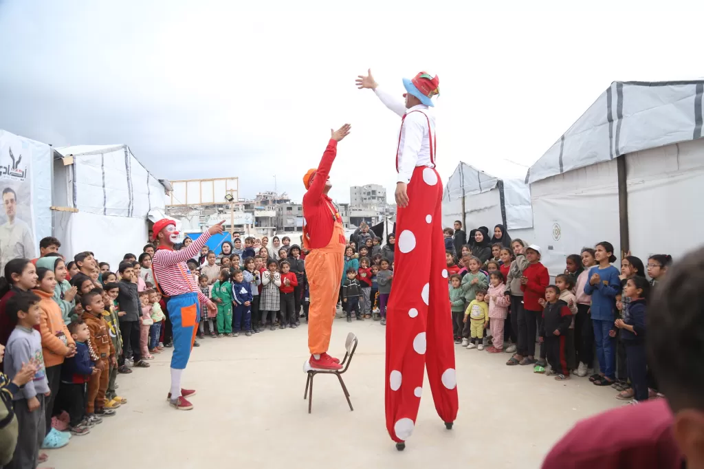 Clowns, including one on stilts, perform for a crowd of smiling children and families in an outdoor camp setting