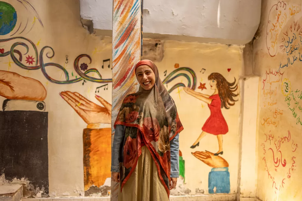 Smiling woman in colorful hijab and shawl standing in front of vibrant wall murals featuring hands, musical notes, and Arabic text