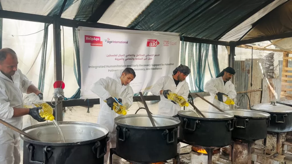 Four men in white aprons cooking in large pots over open fires under a tent with a Age International, HelpAge International, DEC and Chritisian Aid banner