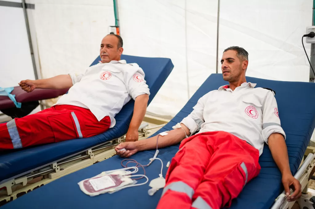 Two paramedics in uniform donating blood on blue medical recliners in a mobile donation unit.
