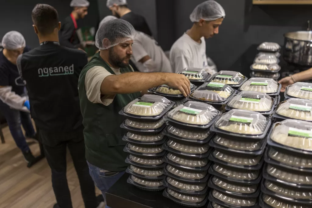 Workers in hair nets and aprons packing prepared meals into black plastic containers in a commercial kitchen or food preparation facility