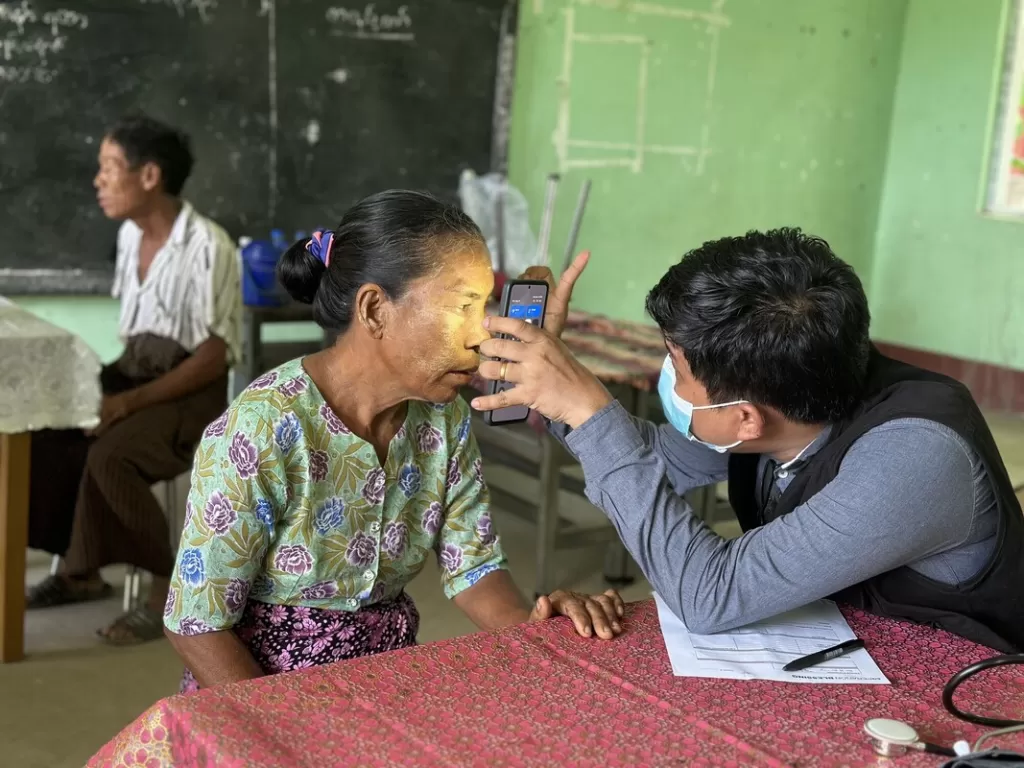 Healthcare worker in mask conducting an eye examination on elderly woman using handheld device in community health clinic.