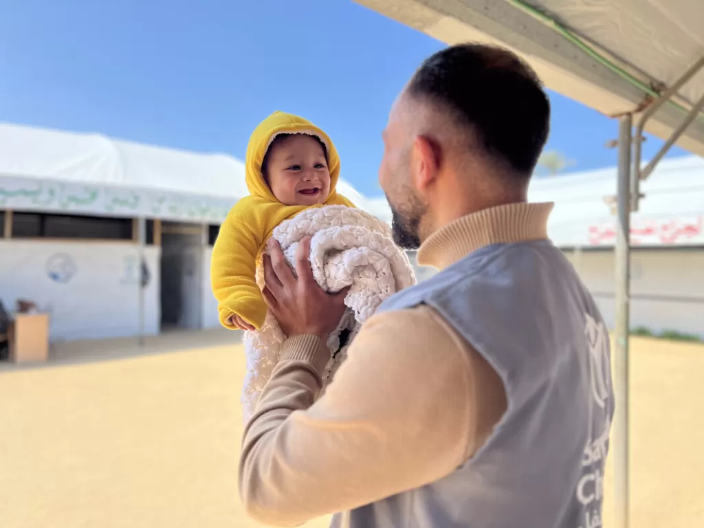 Male aid worker holding smiling baby in yellow hoodie and white blanket outdoors under blue sky with white tent in background