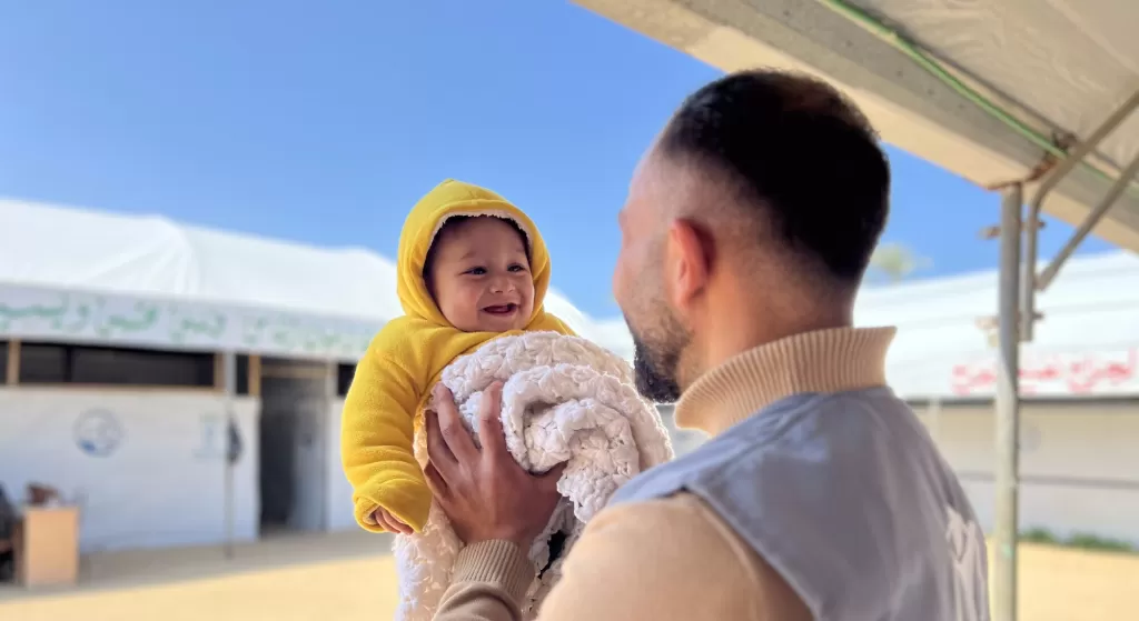Male aid worker holding smiling baby in yellow hoodie and white blanket outdoors under blue sky with white tent in background