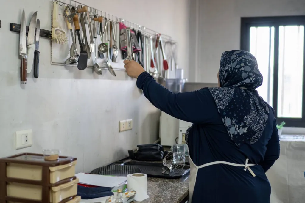 Side and back view of an unidentifiable woman wearing an apron, dark blue top and patterned dark blue headscarf in a kitchen, reaching up to a row of wall-hung cooking utensils