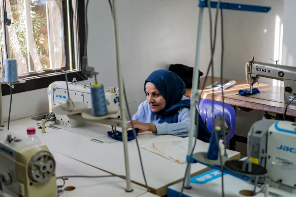 A woman seated at a sewing machine working with other sewing machies and equipment in the room. She wears a blue top and dark blue headscarf 