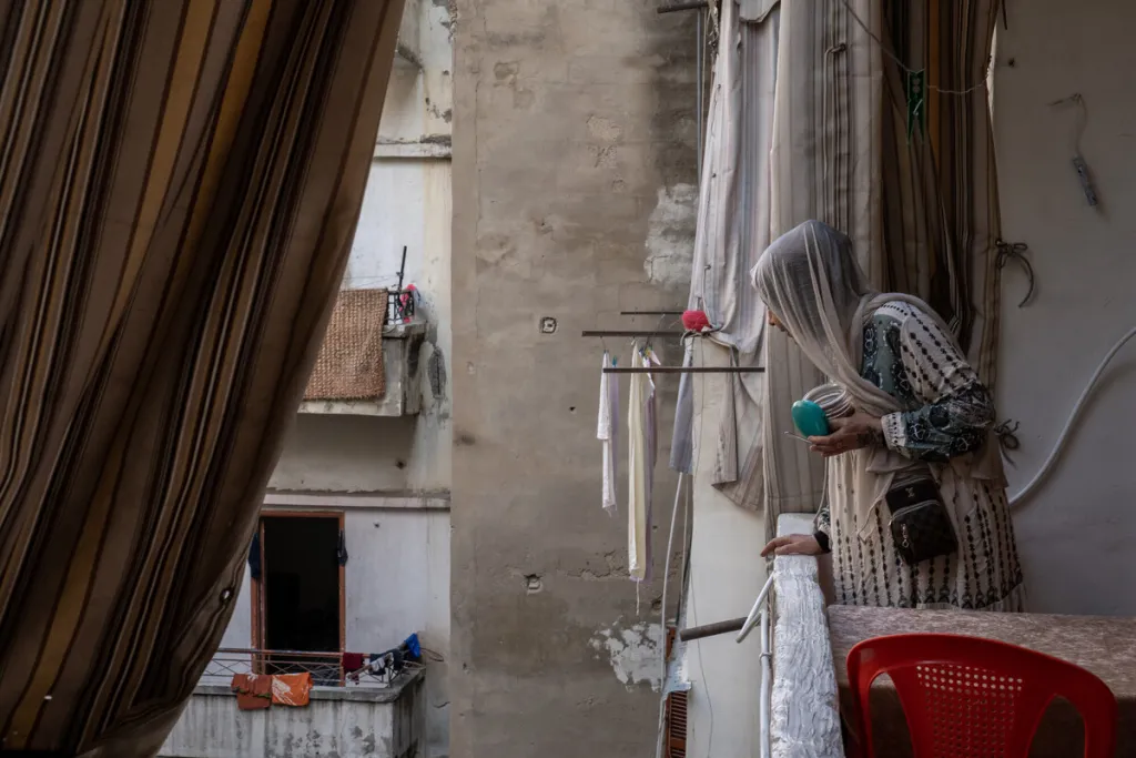 Side view of an unidentifiable woman in a thin white headscarf and loose pale dress peers over a balcony. There is a curtain and red plastic chair in the foreground and clothes drying in the background. 