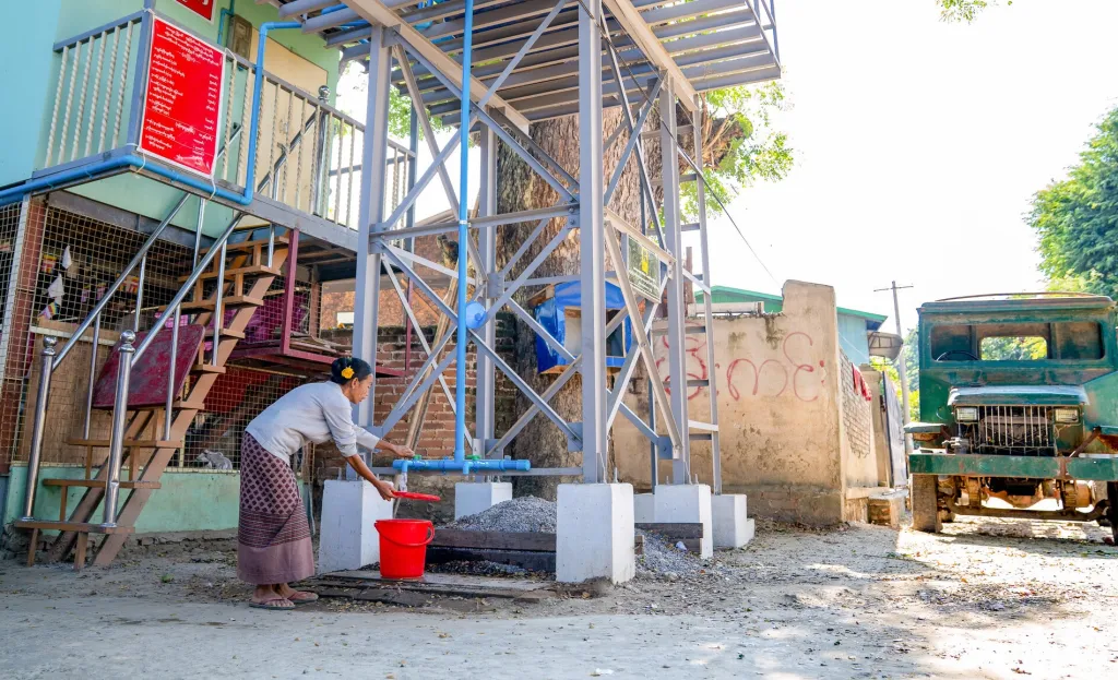 Woman in Myanmar collects water from tap beneath raised metal water tank beside building, filling red bucket in sunny village setting.
