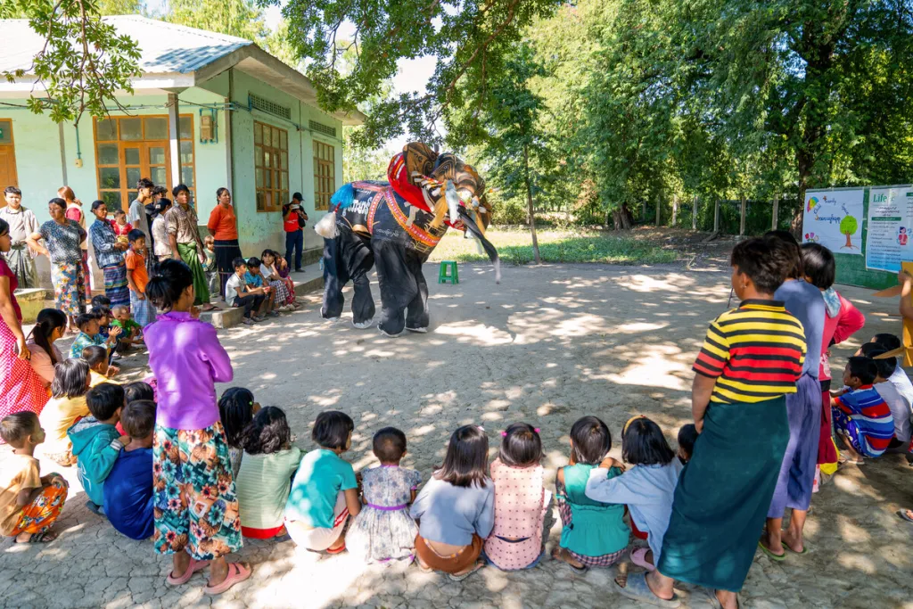 Children and adults in rural Myanmar watch a colourful elephant puppet performance outdoors beside a community building and trees.