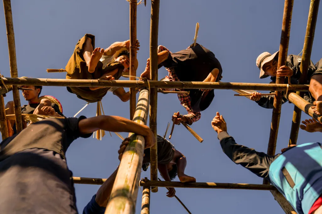 People in rural Myanmar stand on bamboo scaffolding, working together to build or repair a structure under a clear blue sky.