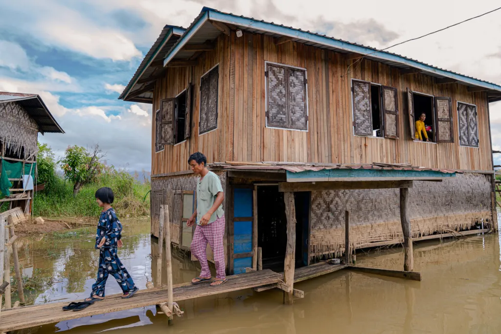 Man and child walk along narrow plank from wooden stilt house surrounded by floodwater in rural Myanmar, while another person watches.