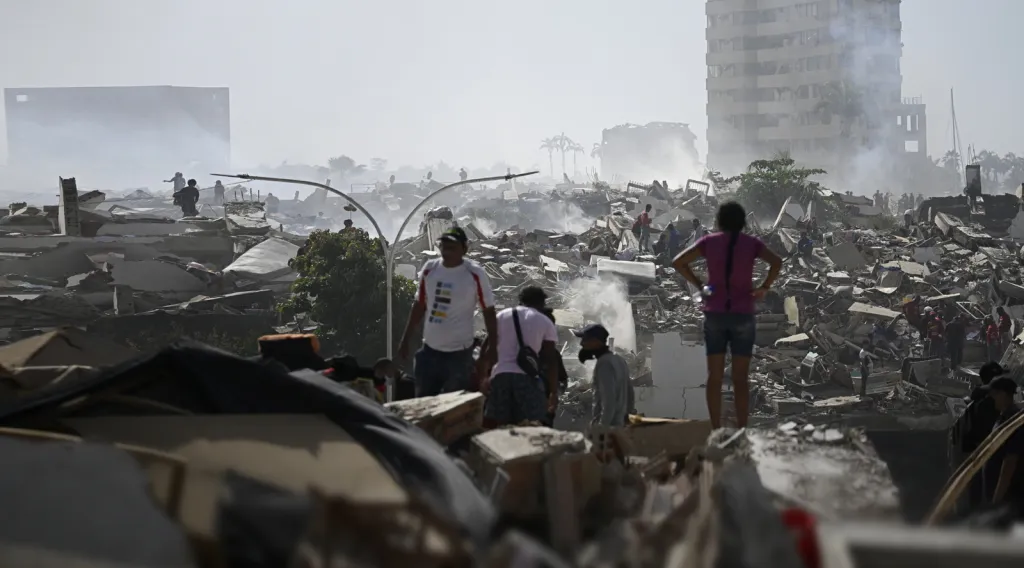 A wide field of concrete rubble with a few remaining buildings in the background amidst the smoke and dust. A few people make their way through the debris, dwarfed by the scale of the destruction.