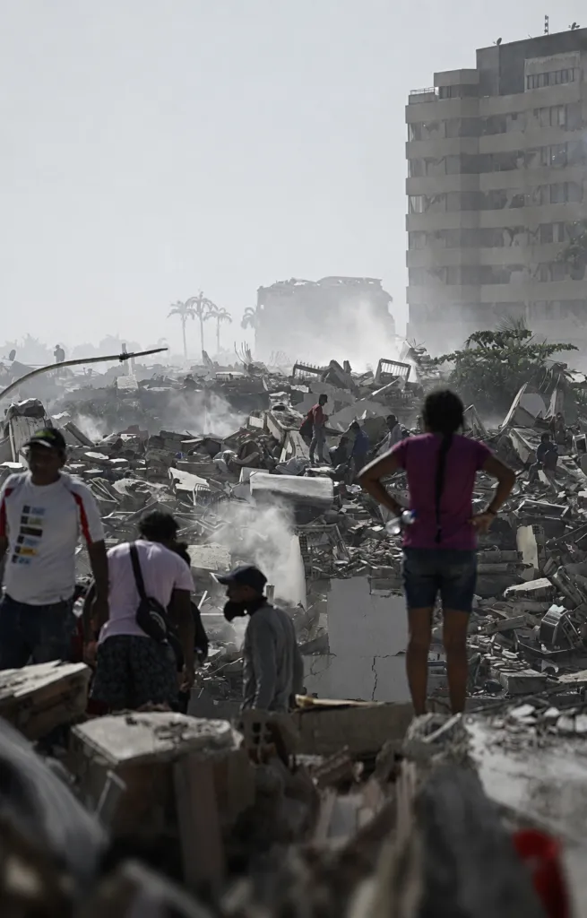 A wide field of concrete rubble with a few remaining buildings in the background amidst the smoke and dust. A few people make their way through the debris, dwarfed by the scale of the destruction.
