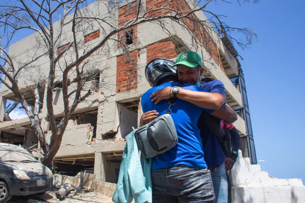 Two men hug in the foreground, visibly upset, with damaged buildings and rubble from the earthquake in the background