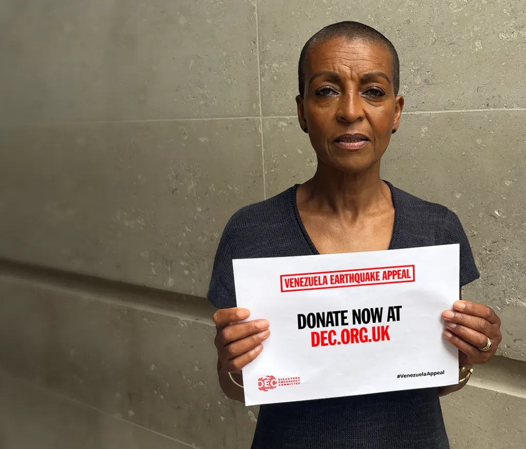 Portrait of Adjoa Andoh in a navy top holding up a paper that says Donate now, dec.org.uk, venezuela Earthquake Appeal, the DEC logo and #VenezuelaAppeal