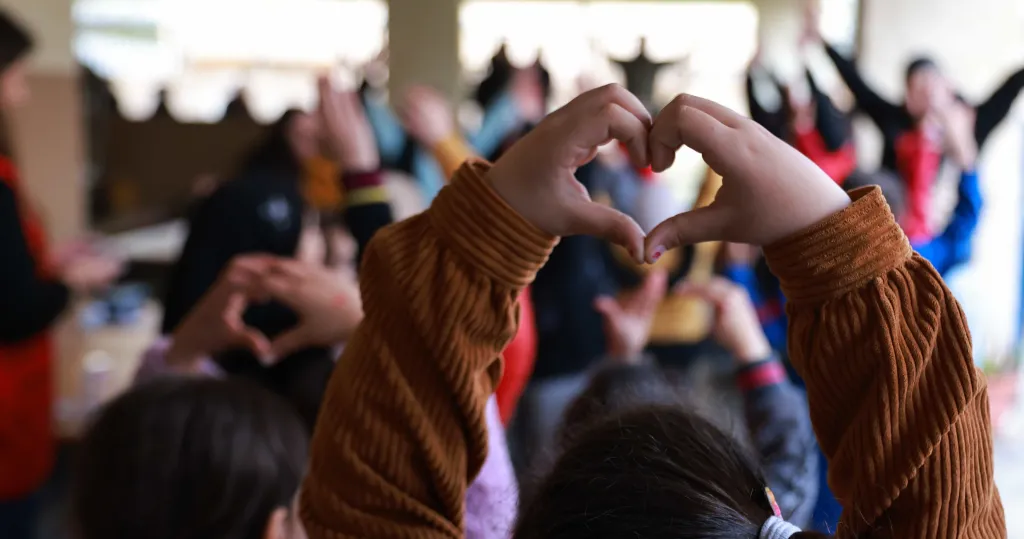 A child makes a heart shape with their raised hands as they stand with a group of other children. Faces are not visible.