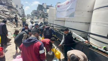 Amidst the rubble, people in Gaza gather to collect water from a water truck as an aid worker fills yellow jerry cans. The truck contains large cyclinders of water and has a banner with the logos of the DEC, member charities and PARC.