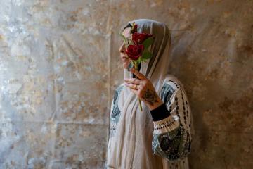 Side view of a woman holding roses that partially block view of her face. The woman is standing against a beige textured background with light coming in from the left. She has a rose tattoo on the hand holding up the dark red flowers. She wears a thin white headcsarf and patterned dress.