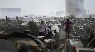 A wide field of heavy concrete rubble is punctuated by a few remaining buildings rising through smoke and dust. A series of human figures pick their way through the debris, dwarfed by the scale of the destruction.