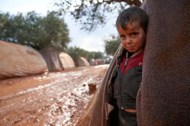 A boy stands at the entrance to a tent in a muddy camp in Syria
