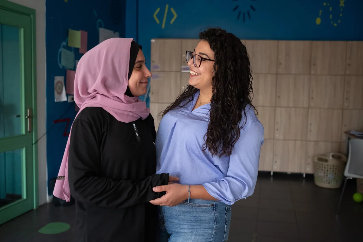 Side view of two women warmly greeting each other and smiling. They are standing in a room with a green door to their right. The woman on the left is wearing a pink headscarf and black loose dress. The woman on the right has glasses, curly hair, wears a pale blue shirt and jeans,